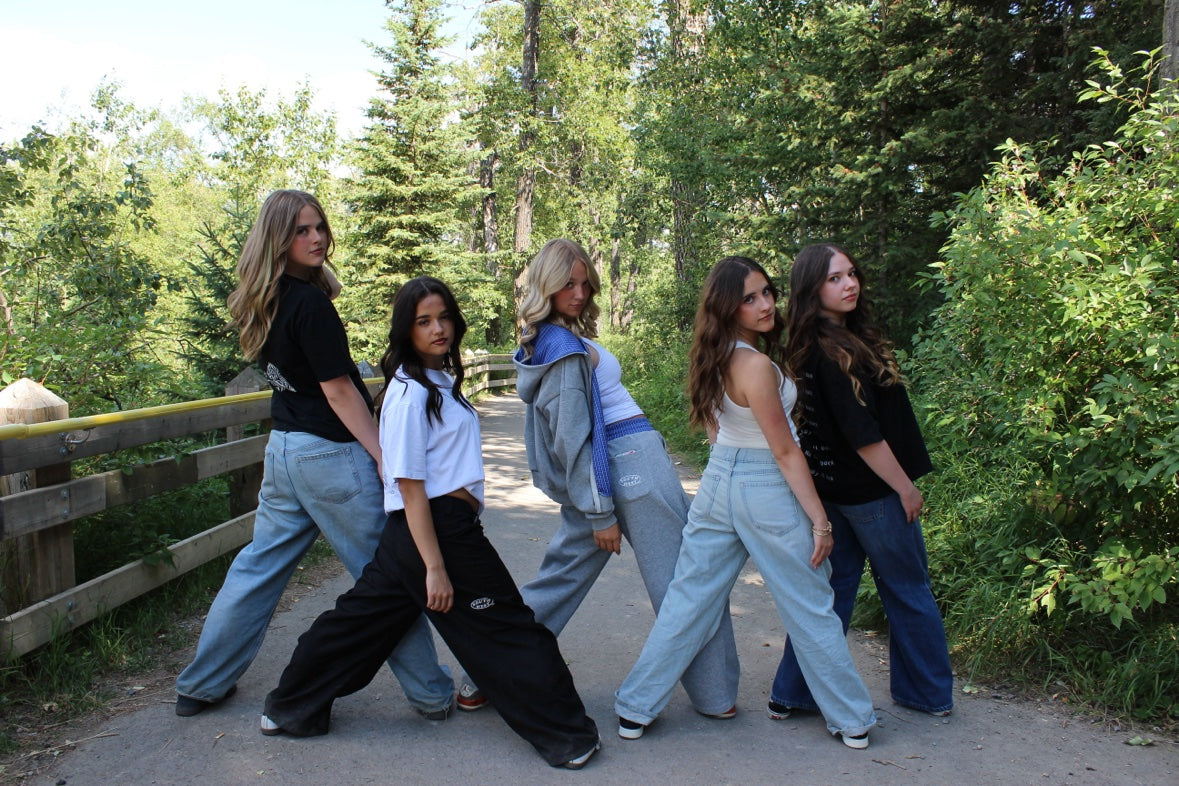 Five young women posing outdoors on a gravel path surrounded by green trees and bushes, wearing various casual streetwear outfits.